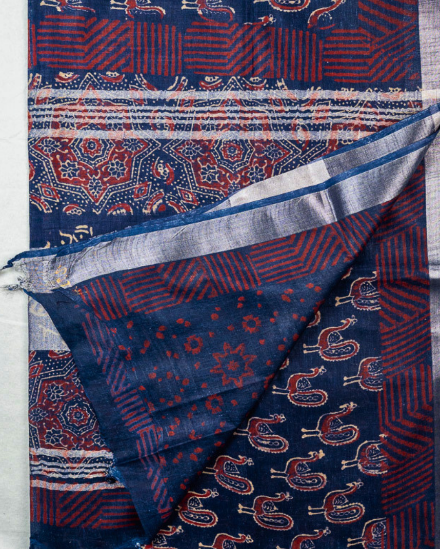 A folded and draped blue patterned cotton shawl or scarf with red and cream motifs, shown against a light background with a brand logo and text at the bottom.